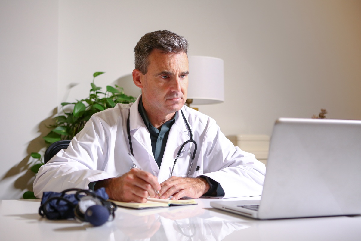 doctor at his desk taking notes from looking at a laptop screen.